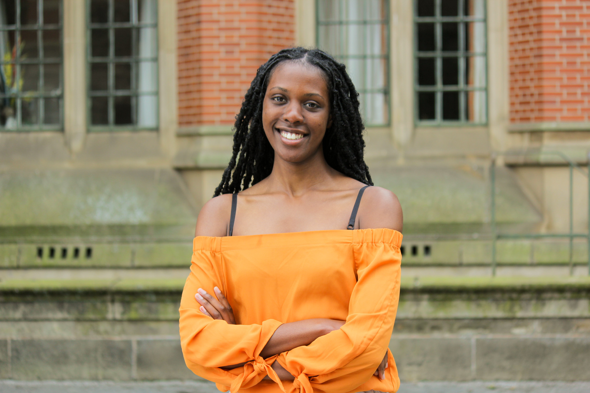 Portrait of Amira Campbell. Amira is smiling into the camera. In the background, the brick wall of a building.